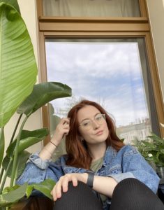 Die Kolumnistin vor einem Fenster sitzend und in die Kamera blickend. Sie hat rotbraune Haare und ein blaues Hemd