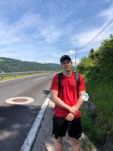 Kolumnist Leon steht beim Wandern an einer Straße, im Hintergrund die Hügel des Alpenvorlands. 