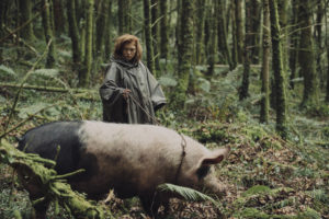 Léa Seydoux aka die Anführerin der Loner führt ein großes Hausschwein durch den Wald. Sie hat rote Haare und trägt ein grünes Regencape.