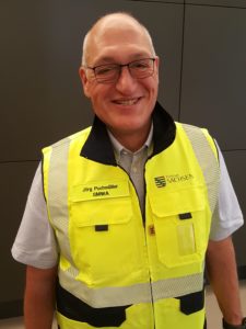Jörg Puchmüller trägt eine gelbe Weste, eine Brille und zeigt ein schönes Lächeln.