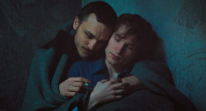 Hans und sein Mithäftling sitzen, sich umarmend, unter einer dünnen grauen Decke nachts in einer Dunklen Ecke, Hans hält eine Zigarette in der Hand.