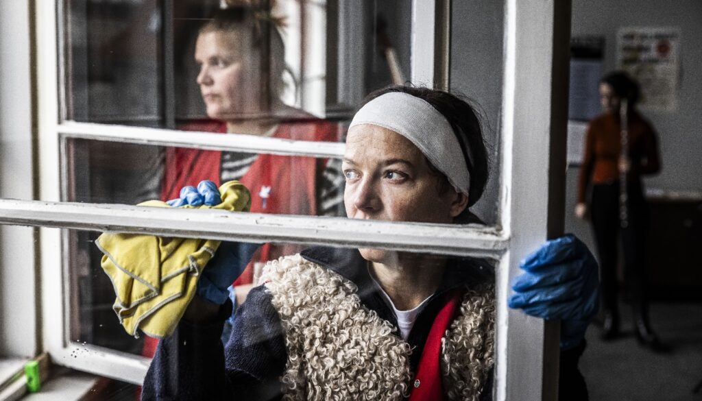 Man sieht die Schauspielerin Hannah Herzsprung in ihrer Rolle als Jenny. Sie putzt gerade ein Fenster und schaut in die Leere. Hinter ihr sieht man zwei Kolleginnen.